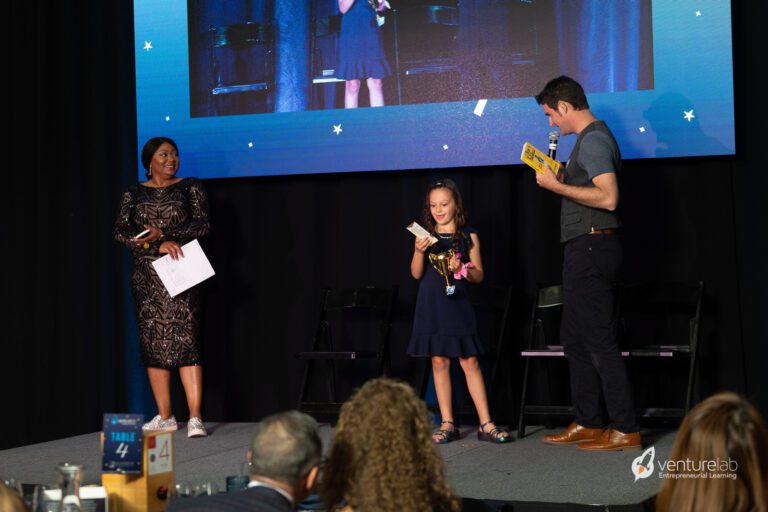 A young girl holding a book and a trophy stands on stage with a man holding a microphone and a woman holding a clipboard, celebrating her achievements in youth entrepreneurship education in front of a large screen displaying their image.