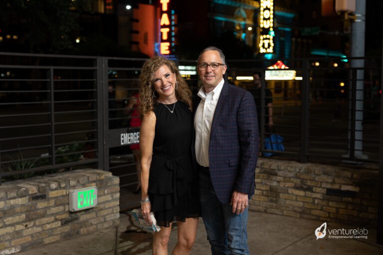 A woman in a black dress and a man in a blue blazer and jeans stand together, smiling, in an outdoor area at night with urban lights in the background. The VentureLab logo, known for promoting youth entrepreneurship education, is visible in the corner.