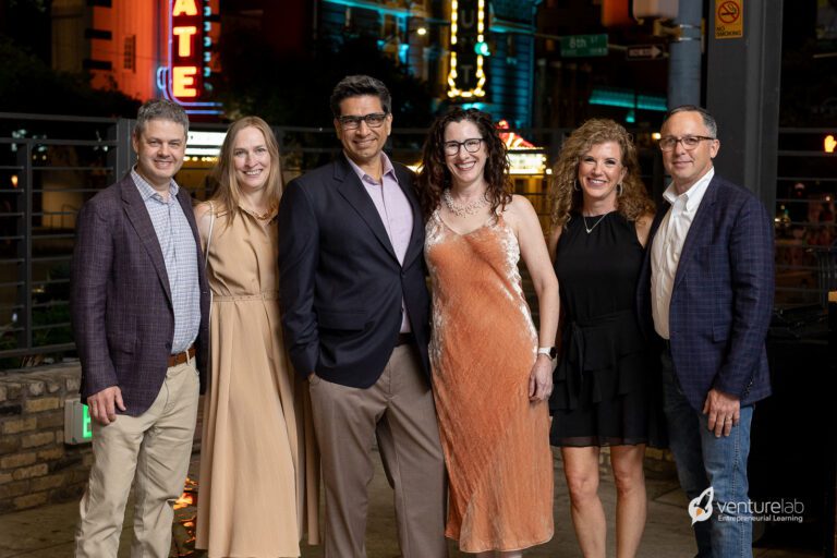 Six people in formal attire are standing in a row, smiling at the camera, with an illuminated city backdrop behind them, celebrating their success in teaching entrepreneurship to young minds.