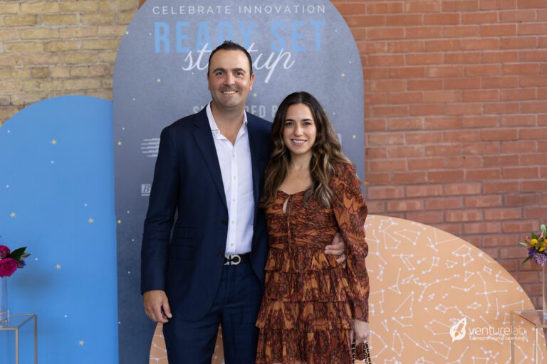 A man in a navy suit and a woman in a patterned brown dress stand together in front of a backdrop that reads 