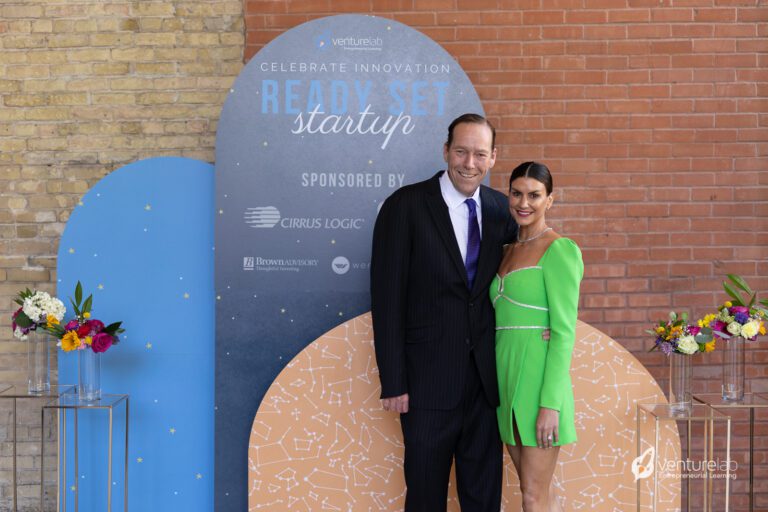 A man in a suit and a woman in a green dress stand together, posing in front of a backdrop for VentureLab's 