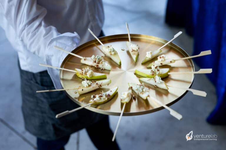 A person holds a gold-colored tray with pear slices topped with cheese and nuts on skewers, reminiscent of the creativity fostered in entrepreneurship education. The person is wearing a white shirt and grey apron, much like a teacher guiding students through a K12 entrepreneur curriculum.