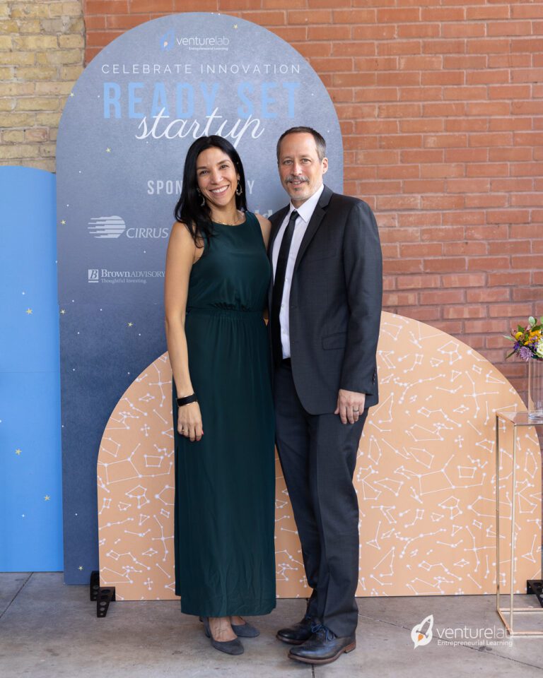 A woman in a green dress and a man in a dark suit pose together in front of a 