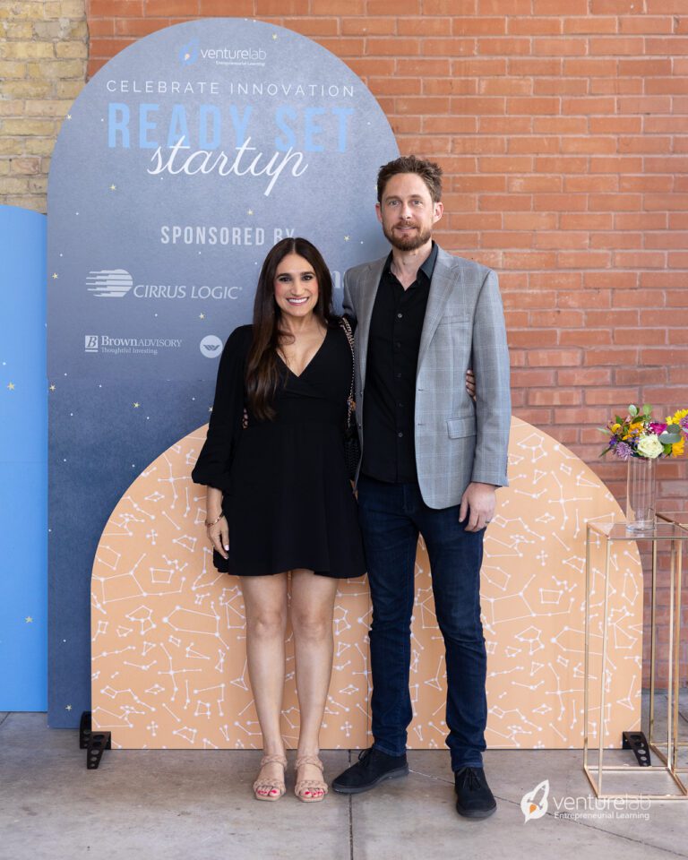 A man and woman stand together in front of a backdrop reading 