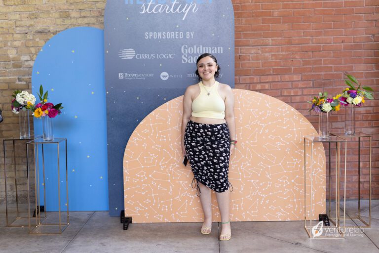 A person stands in front of a decorative backdrop at an event focused on youth entrepreneurship education, wearing a light yellow top and a patterned skirt. There are flower arrangements on stands beside them. Event sponsors' names are visible on the backdrop.