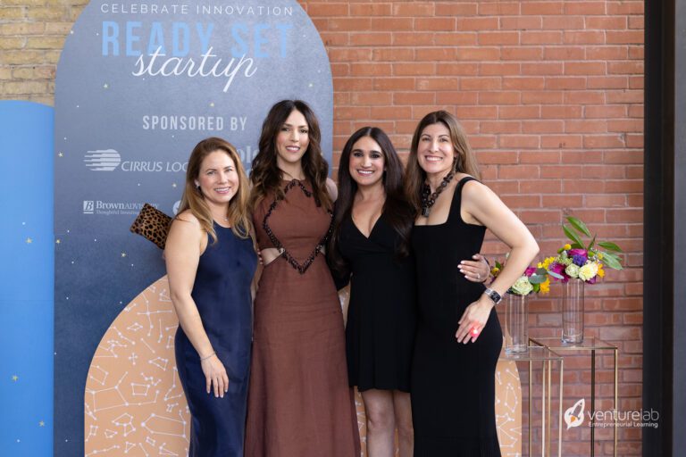 Four women stand together and pose for a photo in front of a sign that reads “Celebrate Innovation: Ready Set Startup.” The event, sponsored by Cirrus Logic, highlights the importance of youth entrepreneurship education. There is a table with colorful flowers.