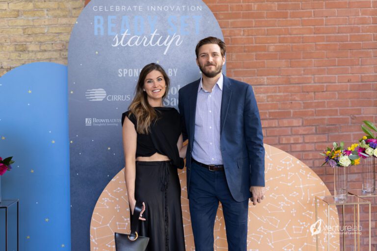 Two people standing and smiling in front of a backdrop with event branding that reads 