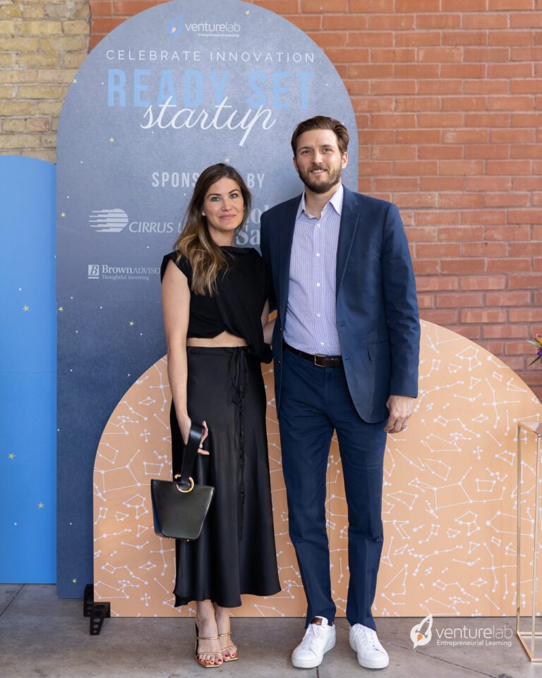 A woman in a black outfit and a man in a blue suit pose together at an event focused on youth entrepreneurship education. VentureLab and sponsors' logos are displayed on a backdrop behind them, highlighting their commitment to teaching entrepreneurship.