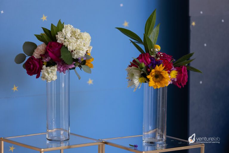Two glass vases with colorful floral arrangements are placed on a glass table against a blue background with star patterns. The vibrant display embodies creativity, much like a K12 entrepreneur curriculum inspiring young minds to innovate and think outside the box.