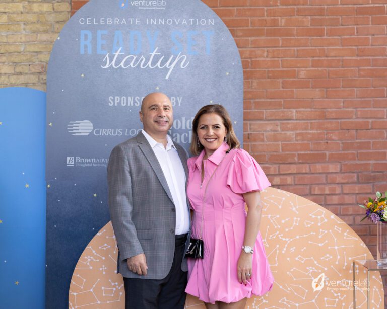 A man in a gray suit and a woman in a pink dress stand together in front of a backdrop with text promoting an event focused on entrepreneurship education.