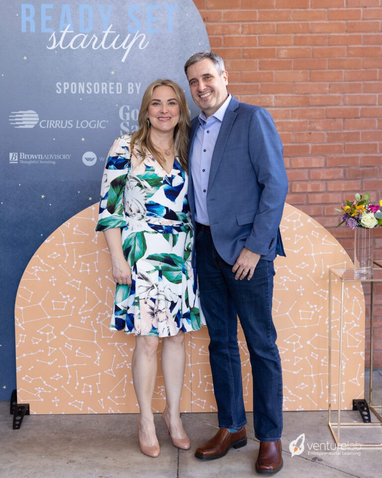 A woman in a floral dress and a man in a blue blazer stand together, smiling, in front of a backdrop with 