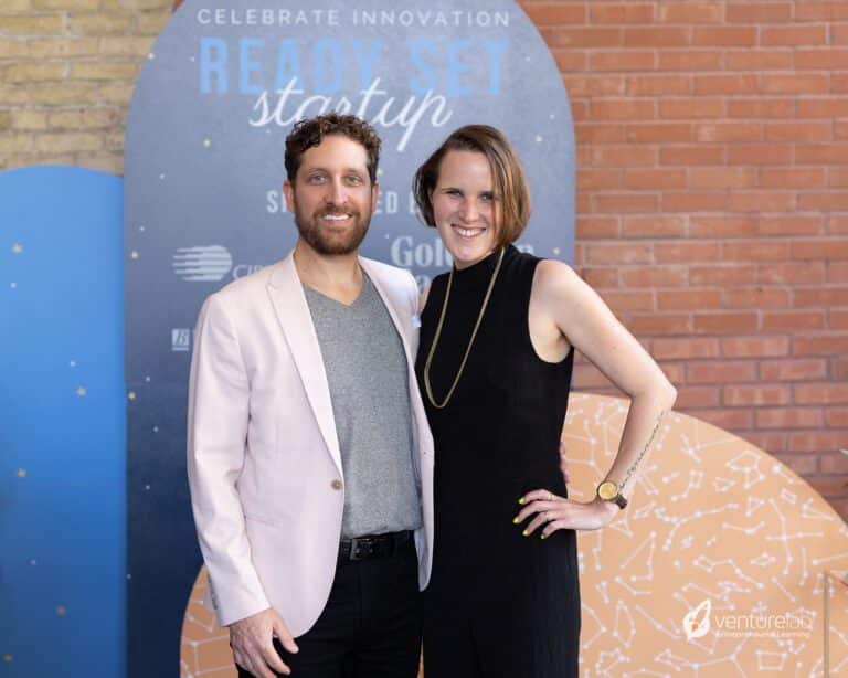 Two people standing together and smiling in front of a backdrop with text 