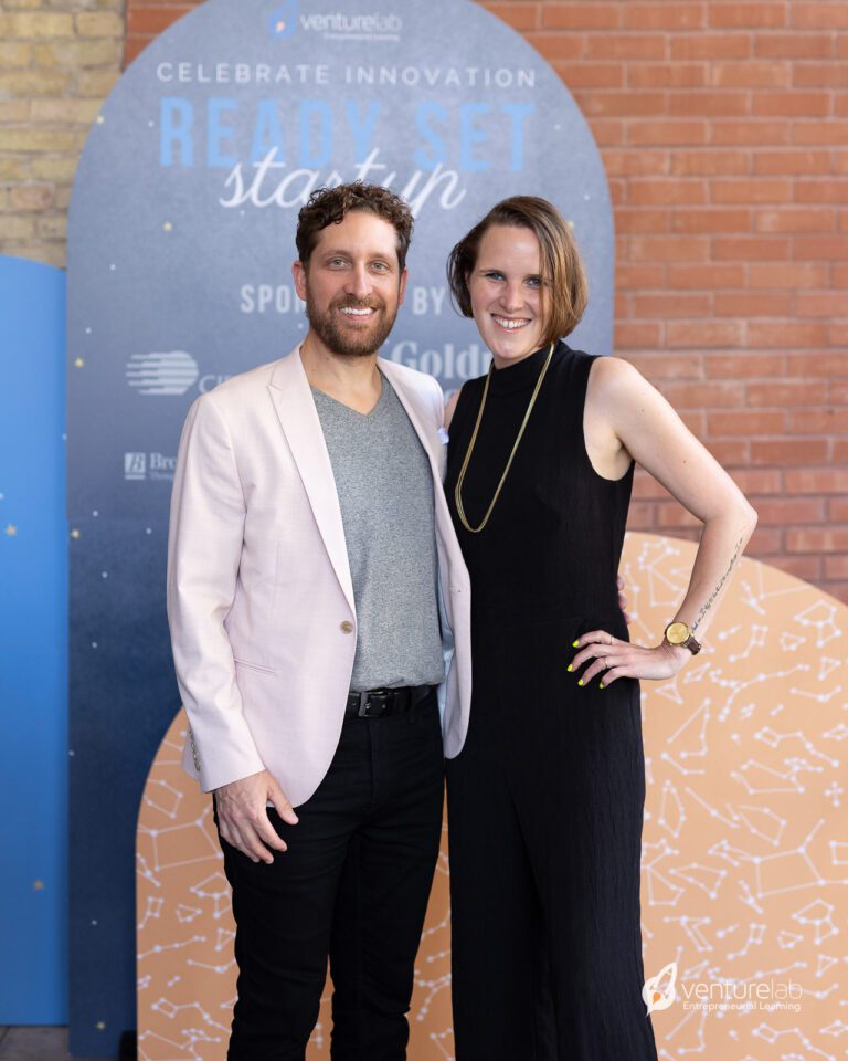 Two people stand side by side at an event focused on youth entrepreneurship education. The man on the left wears a light pink blazer and gray shirt, and the woman on the right is dressed in a black sleeveless outfit. Both are smiling at the camera.