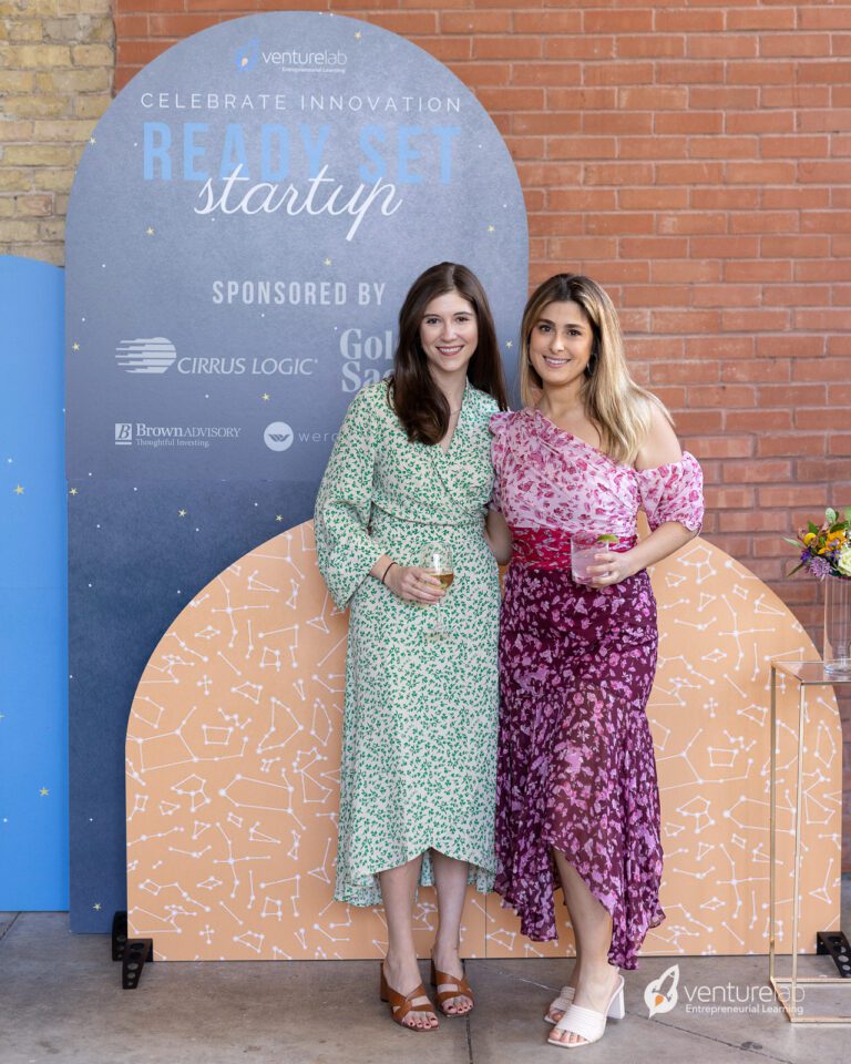 Two women posing and holding drinks in front of a 