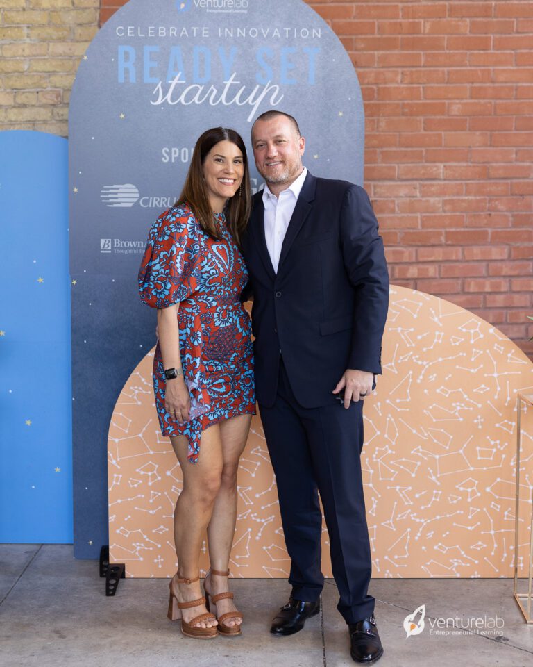 A woman in a patterned blue and orange dress and a man in a dark suit stand together, smiling, in front of a 