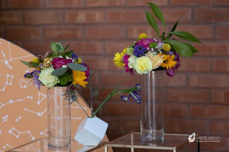 Two flower arrangements in clear glass vases are placed on a glass table against a brick wall backdrop. Both vases contain a mix of colorful flowers and green leaves, reminiscent of the blossoming ideas nurtured through youth entrepreneurship education.