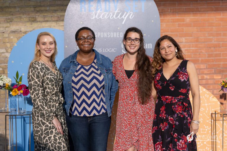 Four women stand side by side, smiling at the camera. They are in front of a display with event branding and sponsor logos, promoting youth entrepreneurship education. Beautiful flower arrangements are placed on either side.