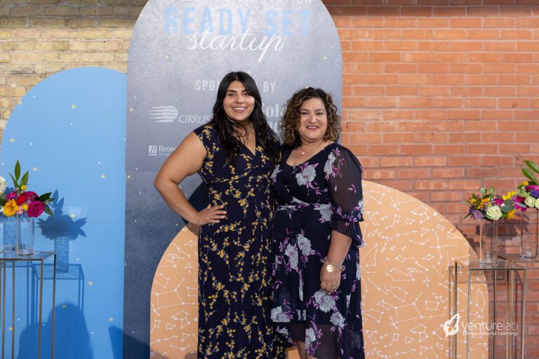 Two women standing side by side, smiling at the camera, in front of a decorated backdrop with text 