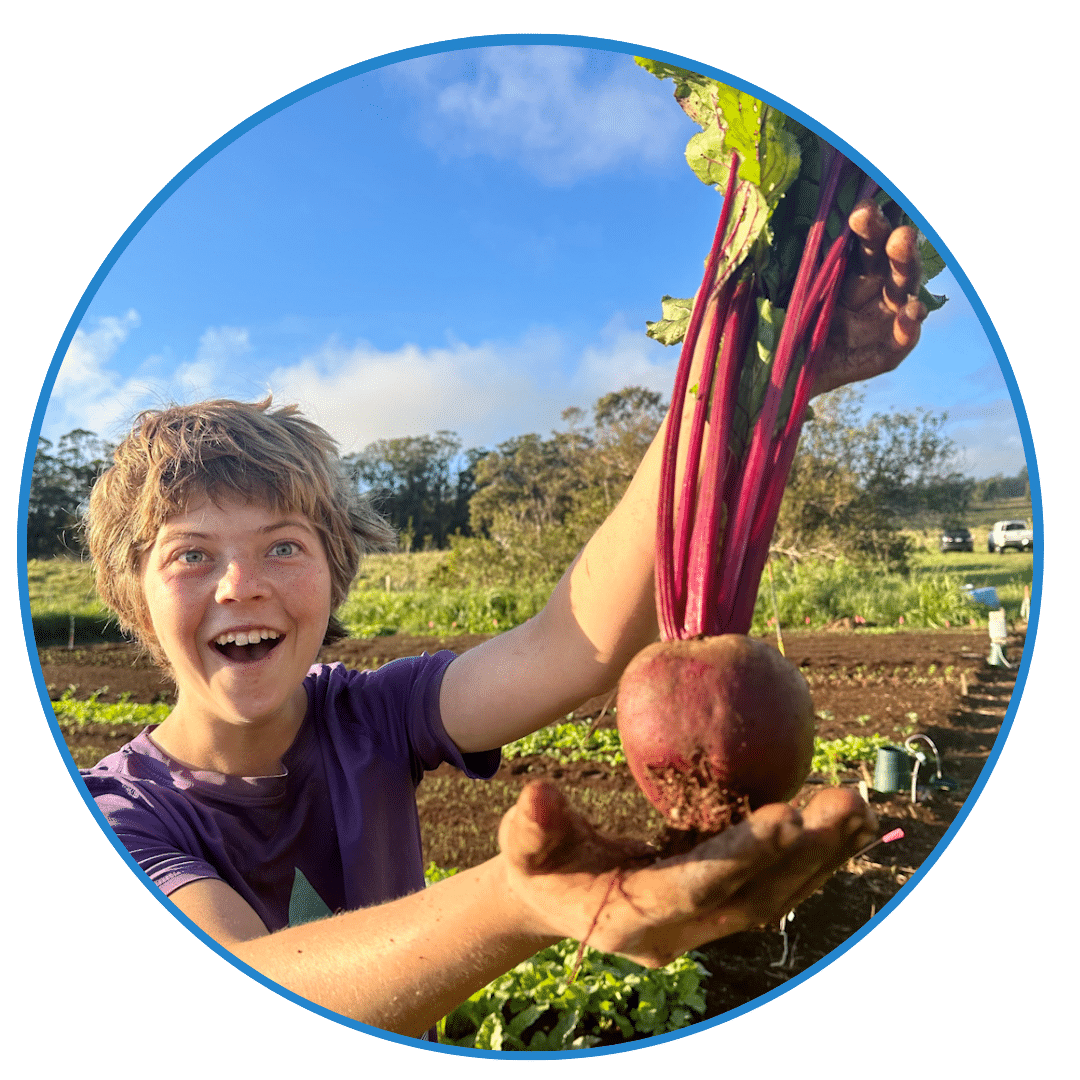 A person in a purple shirt smiles while holding a large beetroot in a garden with greenery and other plants in the background, embodying the hands-on spirit of entrepreneurship education.