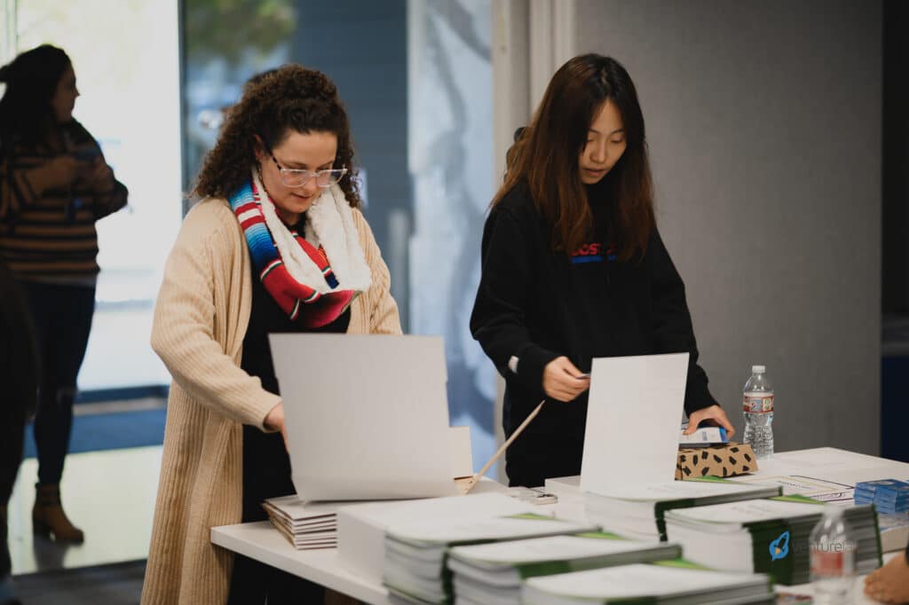 Two women are standing at a table lined with papers and binders. One woman is holding a white paper while the other is leaning over the table, discussing an important aspect of the k12 entrepreneur curriculum. People in the background are blurred.