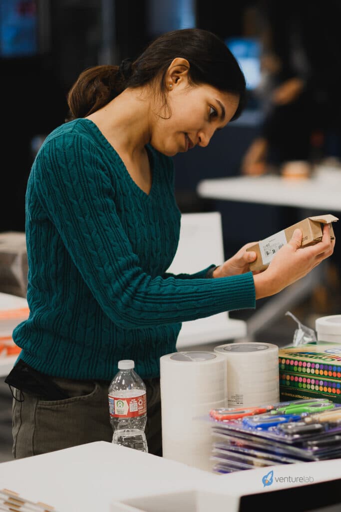 A woman wearing a green sweater attentively examines a small boxed item in her hands, standing near a table with various office supplies and a water bottle, as part of preparing materials for youth entrepreneurship education.