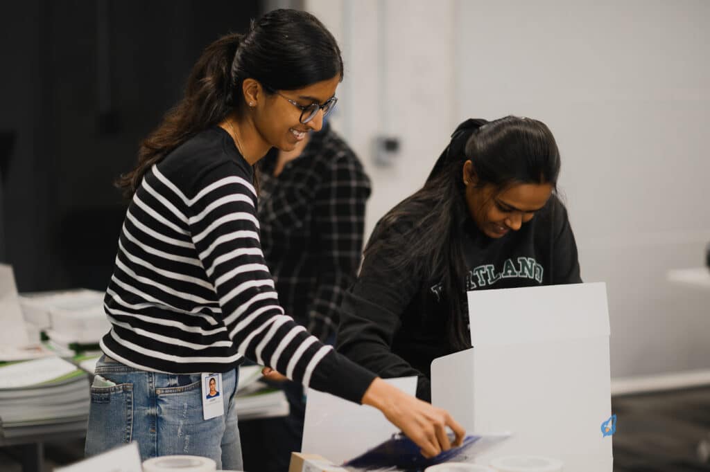 Two people are unpacking or organizing items in a room. One, wearing a striped sweater and glasses, is setting up materials for a k12 entrepreneur curriculum. The other, clad in a sweatshirt, diligently arranges books focused on youth entrepreneurship education. Both appear deeply focused on their tasks.