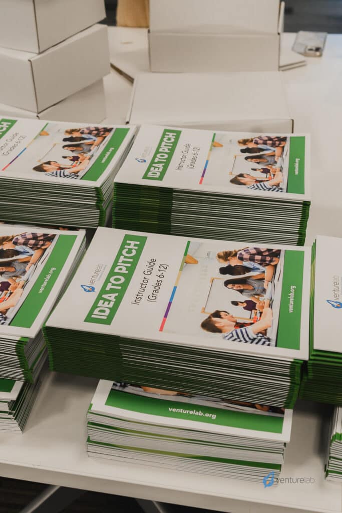 Stacks of "Idea to Pitch: Innovation Guide for Grades 7-12" booklets are neatly arranged on a table. The booklets, part of the k12 entrepreneur curriculum, feature a cover image of students engaging in an activity.
