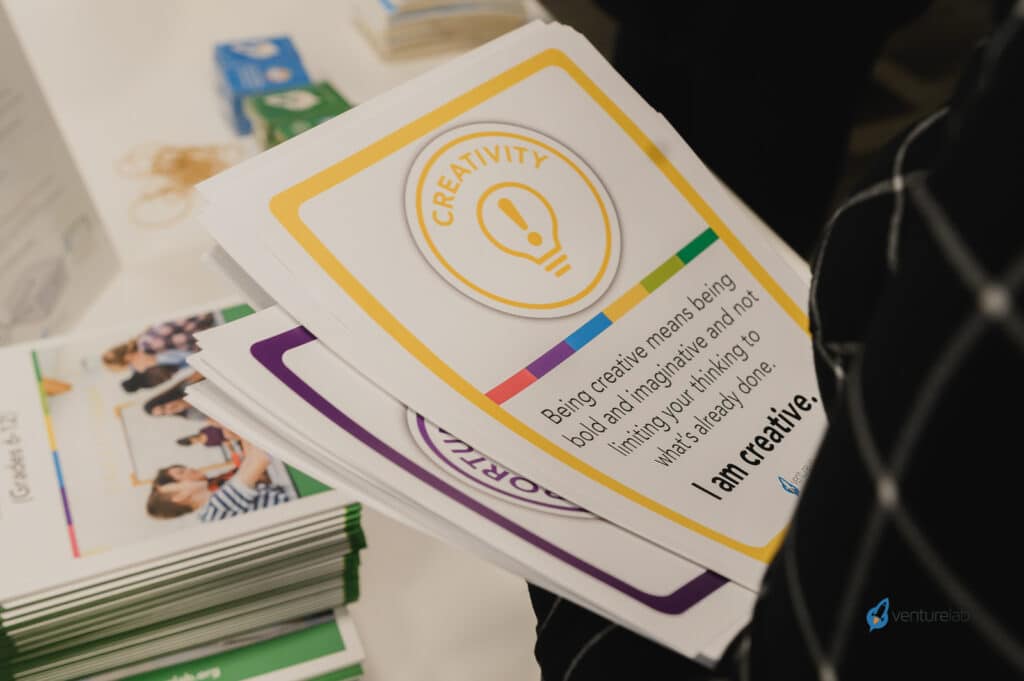 A person holds a paper with a "CREATIVITY" label and text: "Being creative means being bold and imaginative, having no limits in your thinking, always looking for what's not yet thought of—traits vital for youth entrepreneurship education.
