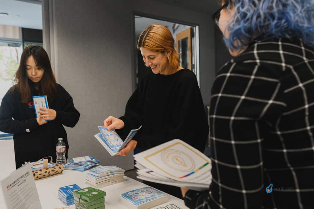 Three people at a table with various pamphlets and booklets; one individual in black is handing out materials on youth entrepreneurship education while two others are looking at items on the table.