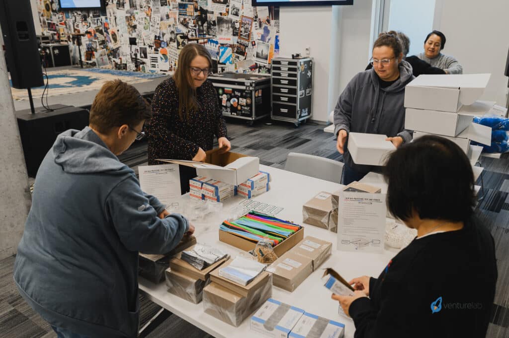 Four individuals are gathered around a table assembling kits with various supplies in a room adorned with posters and screens, all part of an interactive youth entrepreneurship education program.