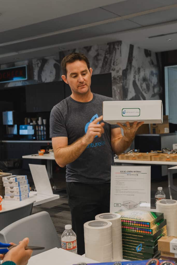 A man in a gray t-shirt is standing in an office, holding a product box and pointing to it. Various items, including bottled water and a stack of boxes, are on the table in front of him. He seems to be involved in teaching entrepreneurship principles through hands-on experience.