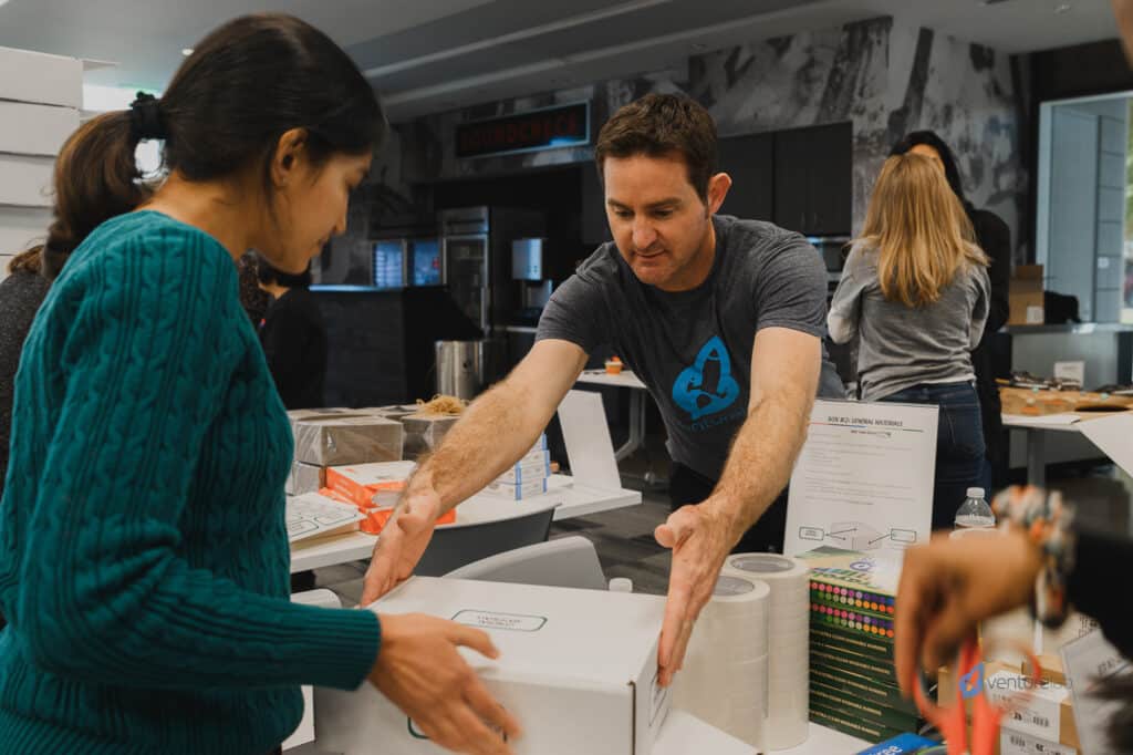 People in a room are focused on packing boxes. A man in a grey shirt hands a box to a woman in a green sweater. Various packing materials and supplies are spread out on the table, an atmosphere that fosters entrepreneurship education.