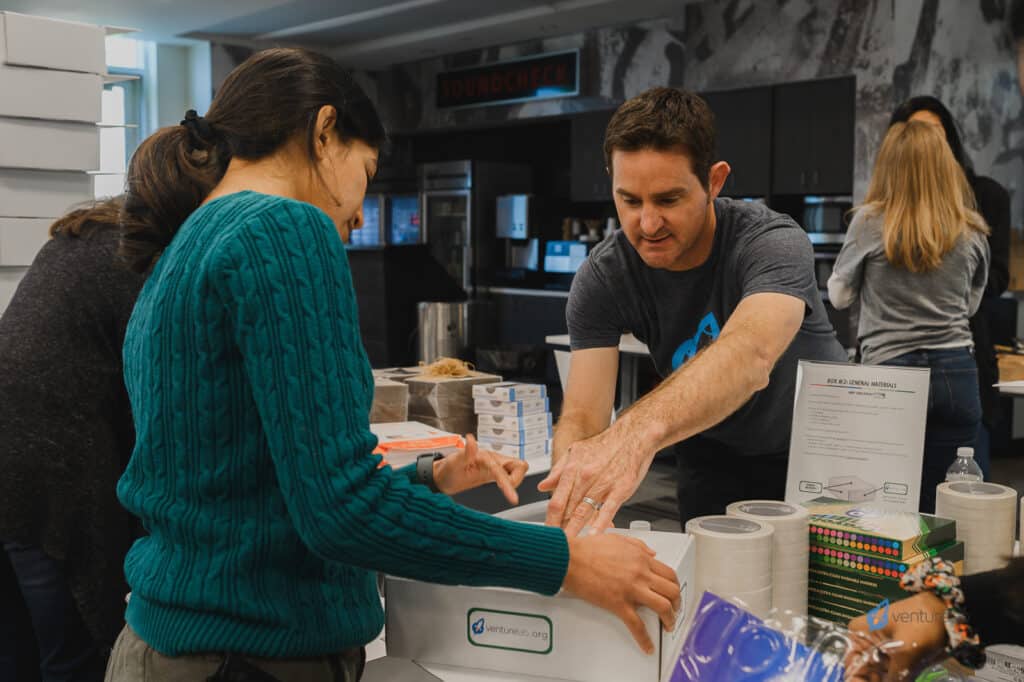 Two people assembling boxes on a table in a room. A man is closing a box, while a woman in a green sweater holds another box. Various packing supplies and tools are around them, demonstrating hands-on activities from the k12 entrepreneur curriculum being taught to eager students. Other people in the background.