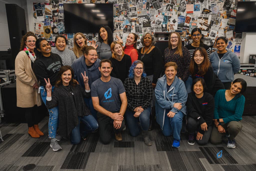 A group of 19 people, consisting of both men and women, pose for a group photo in a room with a collage-covered wall. Several individuals are making playful gestures, embodying the spirit of youth entrepreneurship education.