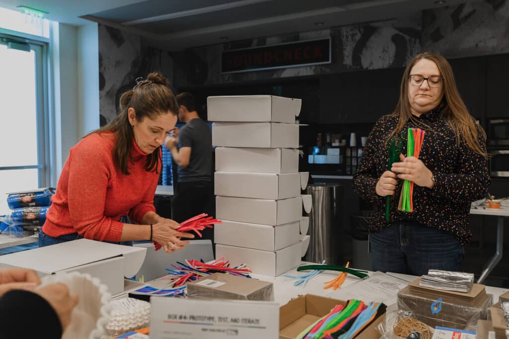 Two women sorting colorful pipe cleaners and stacking white boxes in a room with a sign reading "SOUNDCHECK" in the background, engaging students in hands-on activities as part of their K12 entrepreneur curriculum.