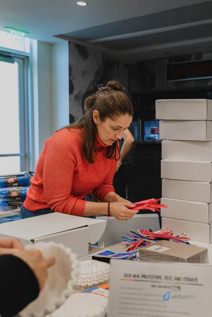 A woman in a red sweater sorts red, white, and blue supplies at a table in a brightly lit room with stacked white boxes and a VentureLab logo on a document, highlighting the importance of entrepreneurship education.