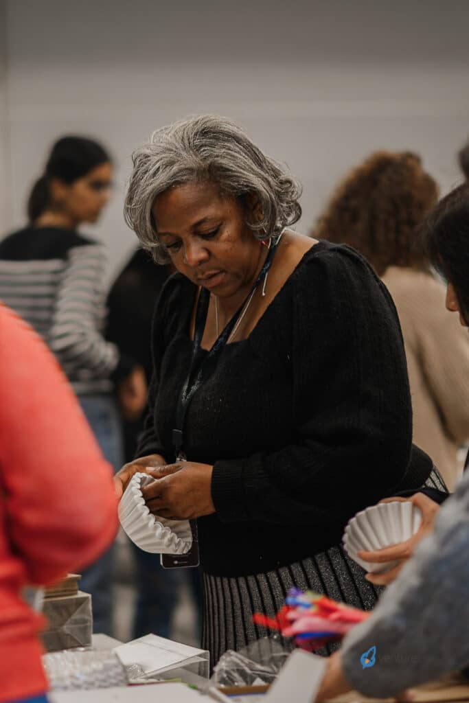 A woman with gray hair and a black sweater is engaged in an activity at a table, holding a white filter. Around her, others are involved in similar tasks as part of a workshop focused on teaching entrepreneurship.