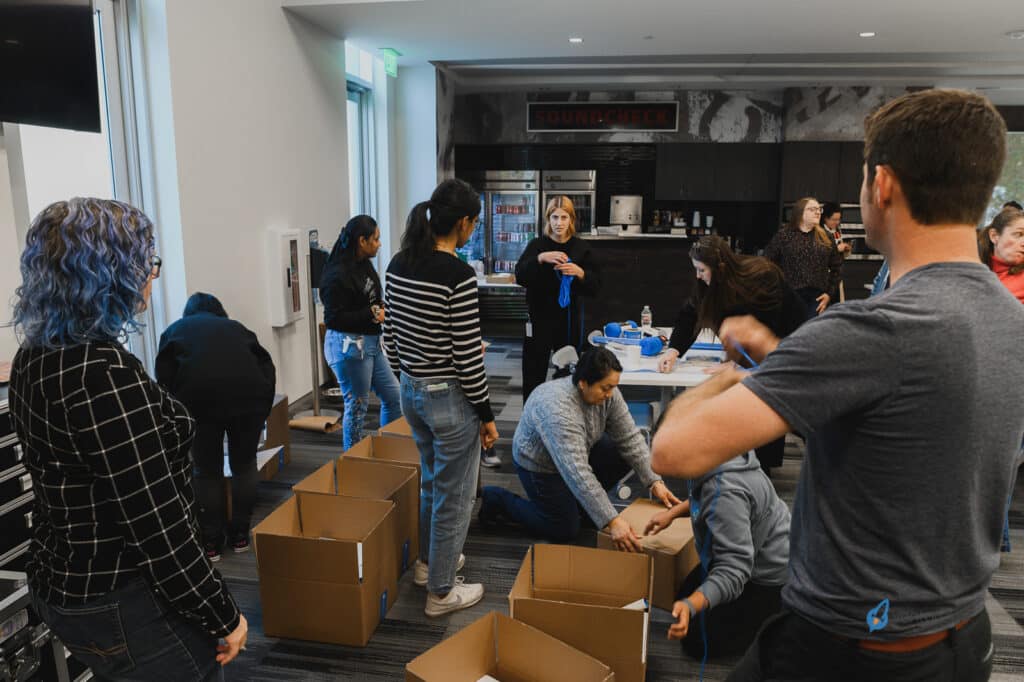 A group of people are assembling boxes in a room. Some are kneeling on the floor handling the boxes, while others stand around engaging in discussion. A woman gestures while speaking at the back, emphasizing points about youth entrepreneurship education through a new K12 entrepreneur curriculum.