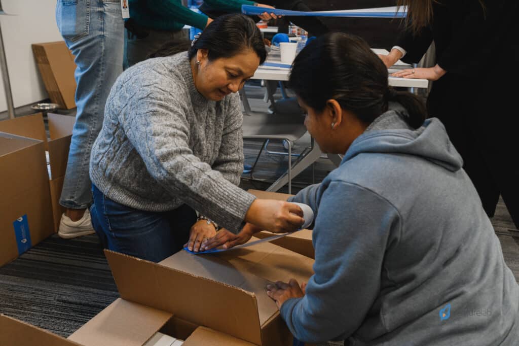 Two individuals are using tape to seal cardboard boxes in an indoor setting. Other people are partially visible, engaged in similar activities, resembling a youth entrepreneurship education class. Apparent teamwork and collaboration in an organized environment.