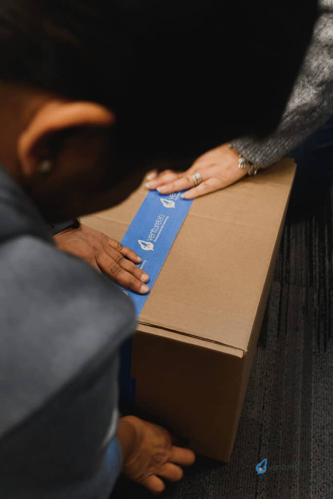 Two individuals are sealing a cardboard box with blue tape that has an "ventures" logo on it, as part of a youth entrepreneurship education initiative.