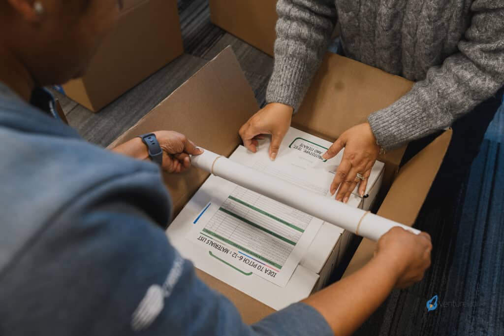 Two people packing documents and a rolled-up chart into a cardboard box, preparing essential materials for a youth entrepreneurship education program.