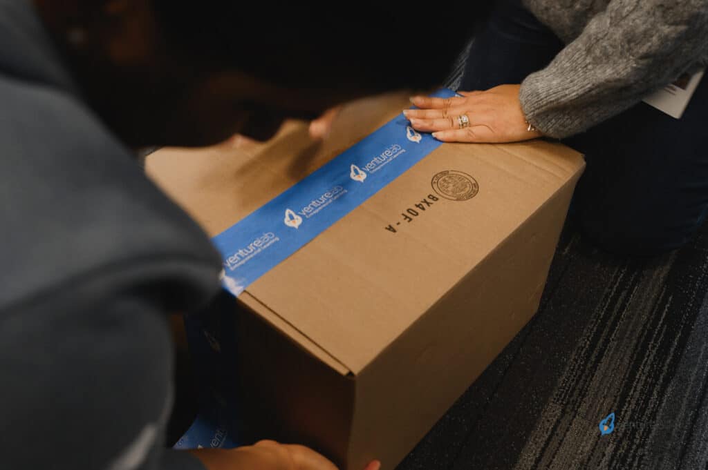 Two people are sealing a brown cardboard box with blue branded tape, demonstrating the principles of youth entrepreneurship education by ensuring the package is secure.