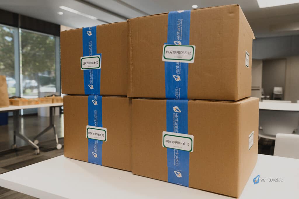 Four sealed cardboard boxes with blue tape labeled "IDEA TO PITCH 6-12" are stacked on a table in an indoor setting, featuring the VentureLab logo on the tape. These tools are essential for teaching youth entrepreneurship education effectively.