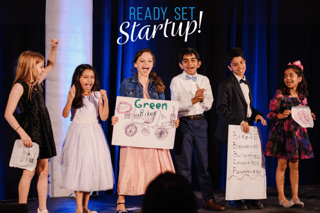 Children presenting business ideas on stage with posters, expressing excitement, under a "ready set startup!" sign at the Youth Entrepreneurship Fundraiser.