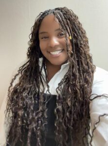 A person with long curly braids smiles at the camera, wearing a white shirt with a black garment over it, proudly showcasing their Springfield High School Entrepreneurship badge against a plain background.