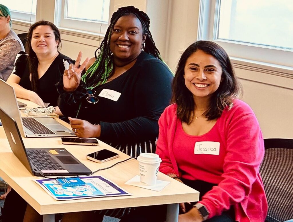 Three people are seated at a table with laptops and papers, smiling at the camera. The person in the middle is making a peace sign. Name tags read "Eboni" and "Jessica." Their enthusiasm reflects a strong commitment to youth entrepreneurship education.