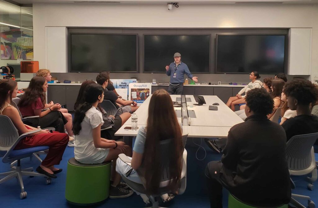 A man standing in front of a group of people sitting around a table at the VentureLab and Saint Louis University partnership meeting.