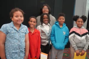 Six children and a woman stand together indoors, all smiling at the camera, exuding confidence that stems from their involvement in a k-12 entrepreneurship curriculum. The children are dressed in casual and semi-formal wear.