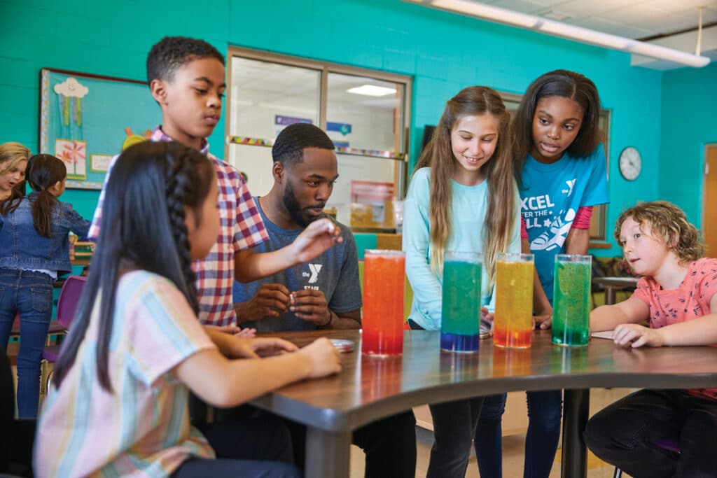 A group of children and an instructor gather around a table with colorful liquid-filled tubes in a classroom setting, showcasing YMCA Chattanooga's VentureLab program that boosts STEM interest with its free entrepreneurship curriculum. Other children interact enthusiastically in the background.