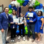 A group of people smiling, with three youths holding awards and certificates from the VentureLab Idea to Pitch program. They stand proudly in front of a colorful balloon arch display.